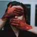 Person with their eyes and mouth covered by another person’s hands smeared in red paint, symbolising silence, control, and violence.