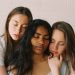 Three young women with their eyes closed gently embrace each other, conveying unity, empathy, and emotional support.